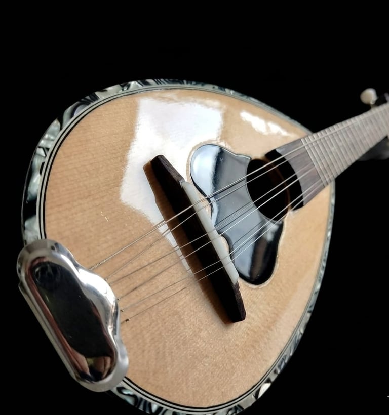 A traditional bouzouki with natural wood body, metal rim, and black pickguard displayed against a black background